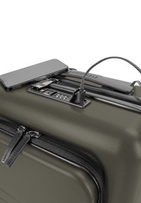 Valigia rigida verde oliva con doppie cerniere nere, lucchetto a combinazione integrato e porta di ricarica. Uno smartphone è appoggiato sopra.