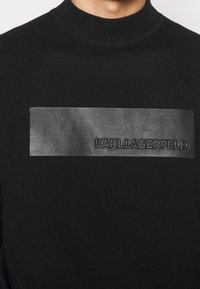 Svart stickad tröja med en framträdande svart läderpatch med "KARL LAGERFELD" präglat. Hög halslinning ger structure.
