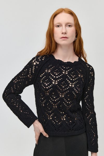 Femme aux cheveux roux et à la peau pâle portant un pull noir en maille ajourée, posant avec une main sur la hanche devant un fond clair uni.