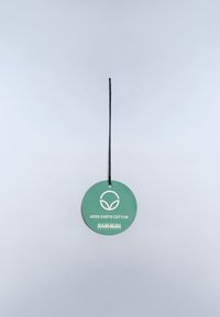 Kruhová zelená visačka na zavesenie s čiernym šnúrkou. Obsahuje logo a text "Good Earth Cotton", s hladkým, matným povrchom.