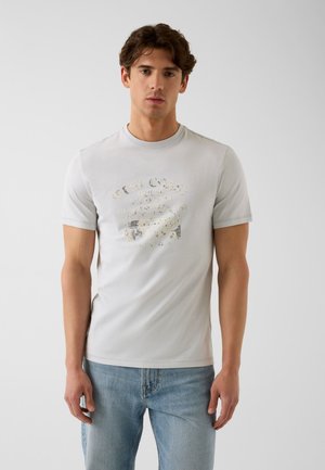 Jeune homme aux cheveux bruns ondulés, portant un t-shirt graphique gris clair et un jean bleu sur un fond blanc uni.