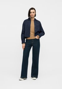 Νεαρή γυναίκα στέκεται με το χέρι στην τσέπη, φορώντας σκούρο μπλε, κοντό σακάκι με κουμπιά, μπεζ ριπ πουλόβερ, τζιν φαρδιάς γραμμής και αθλητικά παπούτσια.