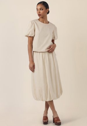 Woman wearing a beige puff-sleeve top and matching gathered midi skirt with brown platform sandals, standing and looking to the side.