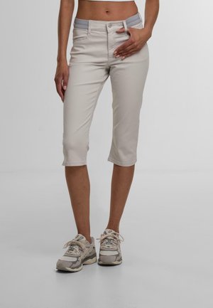 Kvinde iført lyse beige korte bukser og grå sneakers, stående med den ene hånd nær taljen mod en ensfarvet baggrund.