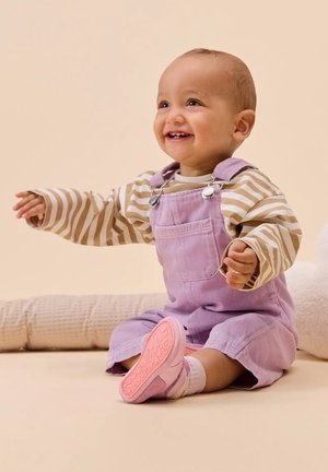 Dunette in corda di lavanda, maglietta a maniche lunghe a righe e scarpe rosa, seduta su una superficie morbida con uno sfondo neutro.
