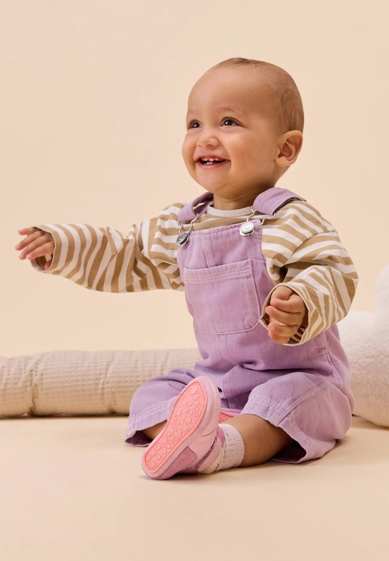 Lavenderfarbene Corduroy-Latzhose, gestreiftes Langarmshirt und rosa Schuhe, sitzend auf einer weichen Oberfläche vor einem neutralen Hintergrund.