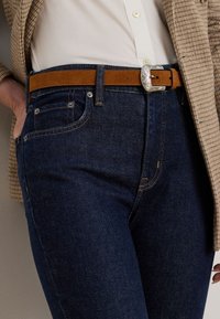 Temno modri jeans z oranžnim šivom, s sprednjimi žepi in svetlo rjavim suede pasom z vpadljivo srebrno sponko.