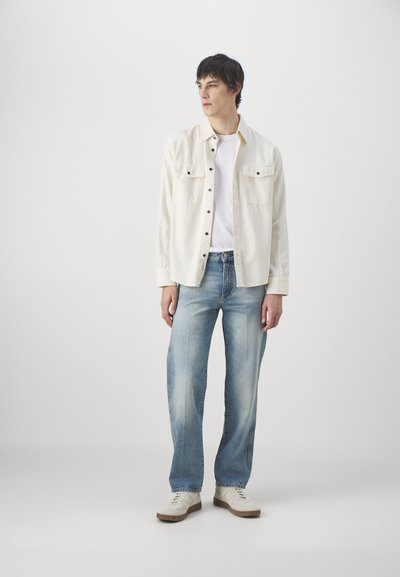 Camisa botón arriba color crema con dos bolsillos en el pecho, llevada sobre una camiseta blanca. Jeans rectos de mezclilla azul claro y zapatillas beige completan el conjunto.