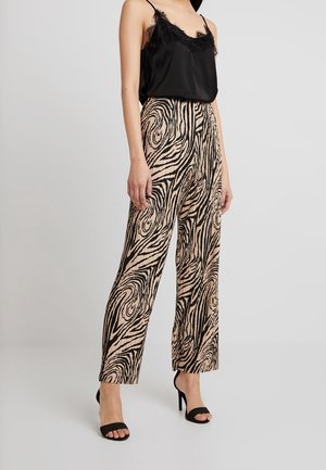 Žena stojící, oblečená v černém tílku s krajkovým lemem, béžových vysokým pasem s černým abstraktním vzorem vířících linií a černých sandálech s otevřenou špičkou.