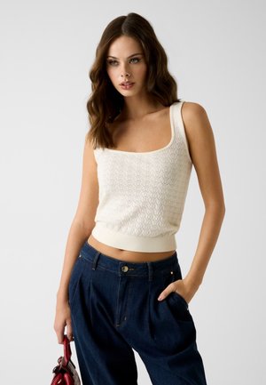 Mujer con cabello castaño ondulado lleva un top de punto blanco sin mangas y vaqueros azul oscuro de talle alto, sosteniendo un pequeño bolso rojo a su lado.