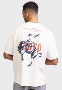 Beżowa koszulka z krótkim rękawem z niebieską grafiką rodeo jeźdźca, z napisem "RODEO" w pogrubionym czerwonym kolorze oraz dodatkowymi napisami u góry.