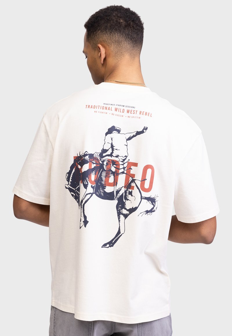 Beżowa koszulka z krótkim rękawem z niebieską grafiką rodeo jeźdźca, z napisem "RODEO" w pogrubionym czerwonym kolorze oraz dodatkowymi napisami u góry.