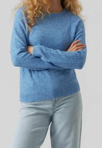 En person klädd i en ljusblå stickad tröja och ljusblå jeans, som står med armarna i kors mot en enfärgad bakgrund.