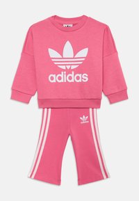 Ensemble de sweat-shirt rose et de pantalons assortis, avec le logo Adidas blanc et trois bandes blanches sur les côtés des pantalons, fabriqué en tissu doux et texturé.