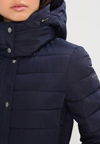 Kobieta w granatowej pikowanej kurtce z wysokim kołnierzem, zatrzaskami i metalowym zapięciem na kapturze.