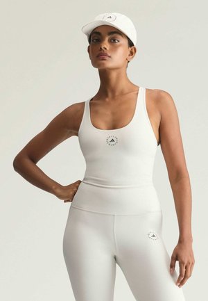 Vrouw in witte atletische tanktop, legging en pet met kleine logo's, staand met één hand op de heup tegen een effen achtergrond.