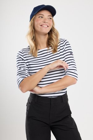 Gestreepte witte en marineblauwe shirt met driekwartmouwen, gecombineerd met zwarte broek. Model draagt een blauwe pet en toont een casual outfit.