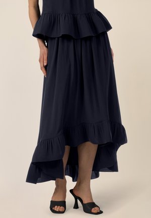 Frau trägt einen marineblauen, gestuften Midirock mit hohem Front- und längerem Rückensaum sowie schwarze, offene Pantoletten mit Absatz, steht vor hellem Hintergrund.