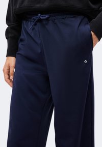 Personne portant un pantalon bleu marine avec cordon de serrage et poches latérales et un haut noir à manches longues, une main dans la poche.
