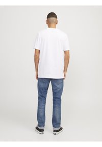 T-shirt en coton blanc à manches courtes, col rond, porté avec un jean en denim bleu et des chaussures noires. L'arrière-plan est uni et de couleur gris clair.
