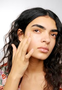 Une personne avec des cheveux bouclés applique un produit léger et crémeux sur sa joue, mettant en valeur une texture de peau naturelle et un subtil éclat.