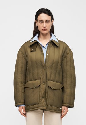 Γυναίκα με φαρδιά μπουφάν από χακί καπιτονέ ύφασμα πάνω από ανοιχτό μπλε πουκάμισο με γιακά, στέκεται μπροστά σε απλό φωτεινό φόντο.