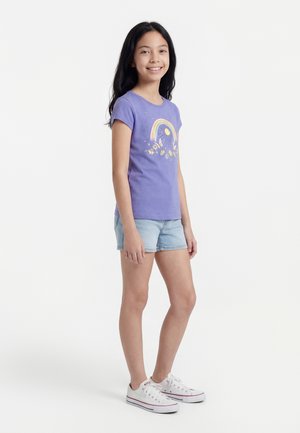 Jeune fille debout, portant un t-shirt violet avec un motif arc-en-ciel et soleil, un short en jean clair, et des baskets blanches sur un fond blanc.