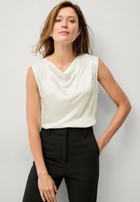 Leichte Cremebluse mit Drapée-Ausschnitt und ärmellosem Design, kombiniert mit maßgeschneiderten schwarzen Hosen und schafft einen professionellen Look.