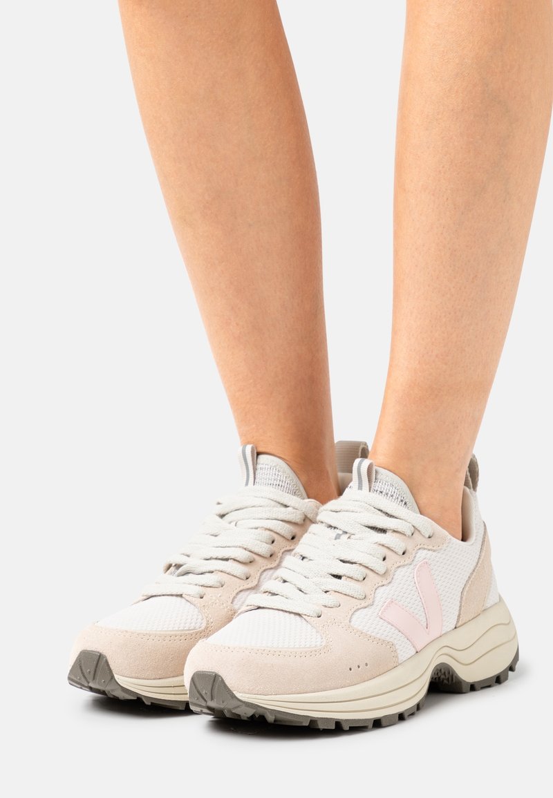 Pieds portant des baskets blanches et rose clair à lacets avec des semelles épaisses texturées sur un fond clair.