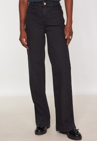 Persona che indossa jeans neri a vita alta, larghi, con tasche a soffietto frontali e mocassini con nappine neri su uno sfondo neutro.