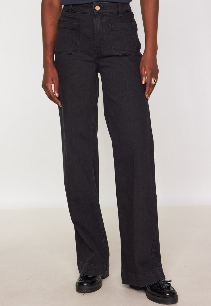 Persona che indossa jeans neri a vita alta, larghi, con tasche a soffietto frontali e mocassini con nappine neri su uno sfondo neutro.