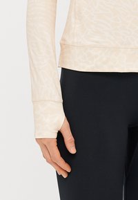 Helebeeživ värvi pika varrukaga topp, millel on peen muster ja mis on valmistatud siledast kangast. Omab kinniseid mansette, mis on varrukate jaoks avatud ning on paaritatud mustade retuusidega.