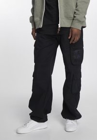 Černé cargo kalhoty s více bočními kapsami, rovný střih, odolná tkanina a jemné logo; doplněné bílými teniskami.