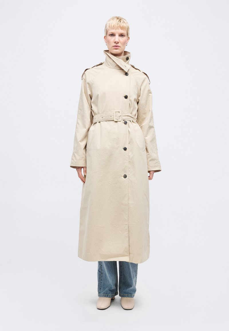 Άτομο που στέκεται φορώντας μια μπεζ μακριά παλτό τύπου trench με ζώνη πάνω από μπλε τζιν και μπεζ παπούτσια, μπροστά σε απλό φωτεινό φόντο.