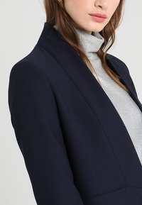 Marinblå blazer med skräddarsydd passform, med en slät yta och sjalkrage; b ärs över en tight passande metallisk silverfärgad polotröja.