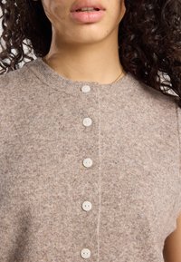 Cardigan en tricot de couleur marron clair avec une texture côtelée, encolure ronde et cinq boutons blancs à l'avant.