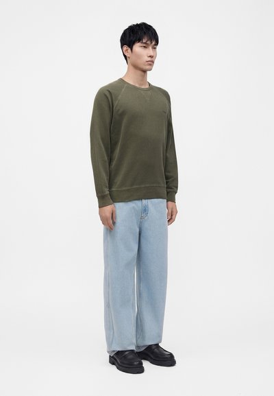 Homme debout face à l'avant portant une chemise à manches longues vert olive, un jean bleu clair à jambes larges et des chaussures noires sur fond blanc.