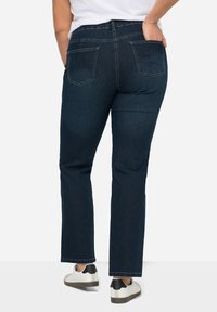 Tamsiai mėlynos džinsinės kelnės su tiesiu kirpimu, turinčios dvi užpakalines kišenes ir subtilius siuvinėjimo elementus. Dėvimos su baltais sportiniais bateliais.