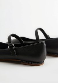 Svarta läderballetskor med rund tå, ankelrem och texturerad svart detaljering på sulan. Slät yta, minimalistisk design.