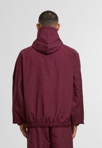 Bordowa pikowana kurtka z kapturem, z elastycznymi mankietami i dołem. Teksturowana powierzchnia z okrągłymi wzorami dodaje szczegółów. Widok z tyłu.