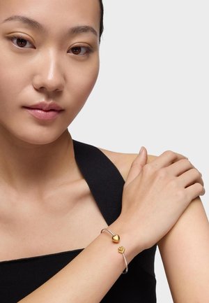 Młoda kobieta w czarnym topie nosząca srebrną bransoletkę mankietową z zawieszkami w kształcie złotego serca i korony na nadgarstku na jednolitym tle.