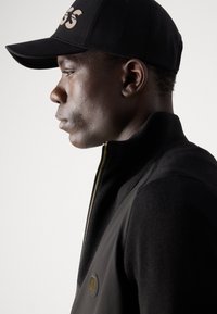 Jachetă neagră cu fermoar, cu guler tricotat și țesătură texturată, asociată cu o șapcă neagră ce are un logo proeminent pe față.