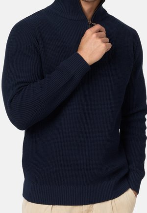 Homme portant un pull en maille texturée bleu marine foncé avec une fermeture éclair au quart, tirant la fermeture éclair vers le haut, pantalon beige visible à la taille.