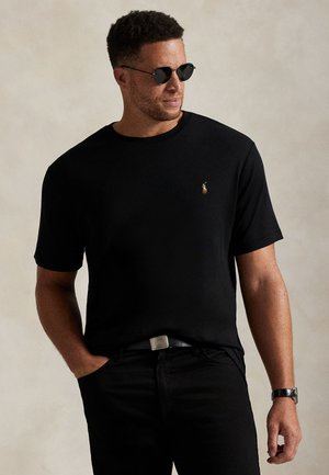 Homme aux cheveux courts et bouclés portant des lunettes de soleil noires, un t-shirt noir avec un petit logo, un pantalon noir et une montre, debout avec une main dans la poche.