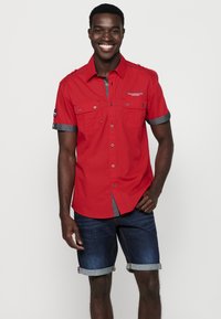 Camisa de botones de manga corta roja con dos bolsillos frontales, acentos de mezclilla en las mangas, combinada con pantalones cortos de mezclilla doblados. Textura suave.