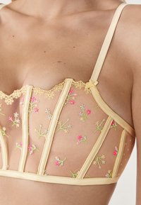 Gelbes Mesh-Bralette mit floraler Stickerei in Pink und Grün, mit gewelltem Spitzenbesatz und dünnen Trägern. Halbtransparentes Stoffmaterial.