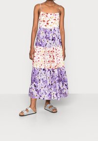 Femme vêtue d'une robe sans manches à volants florale dans des tons de violet et blanc, avec des fines bretelles et des sandales à boucle argentée, se tenant sur un fond uni.