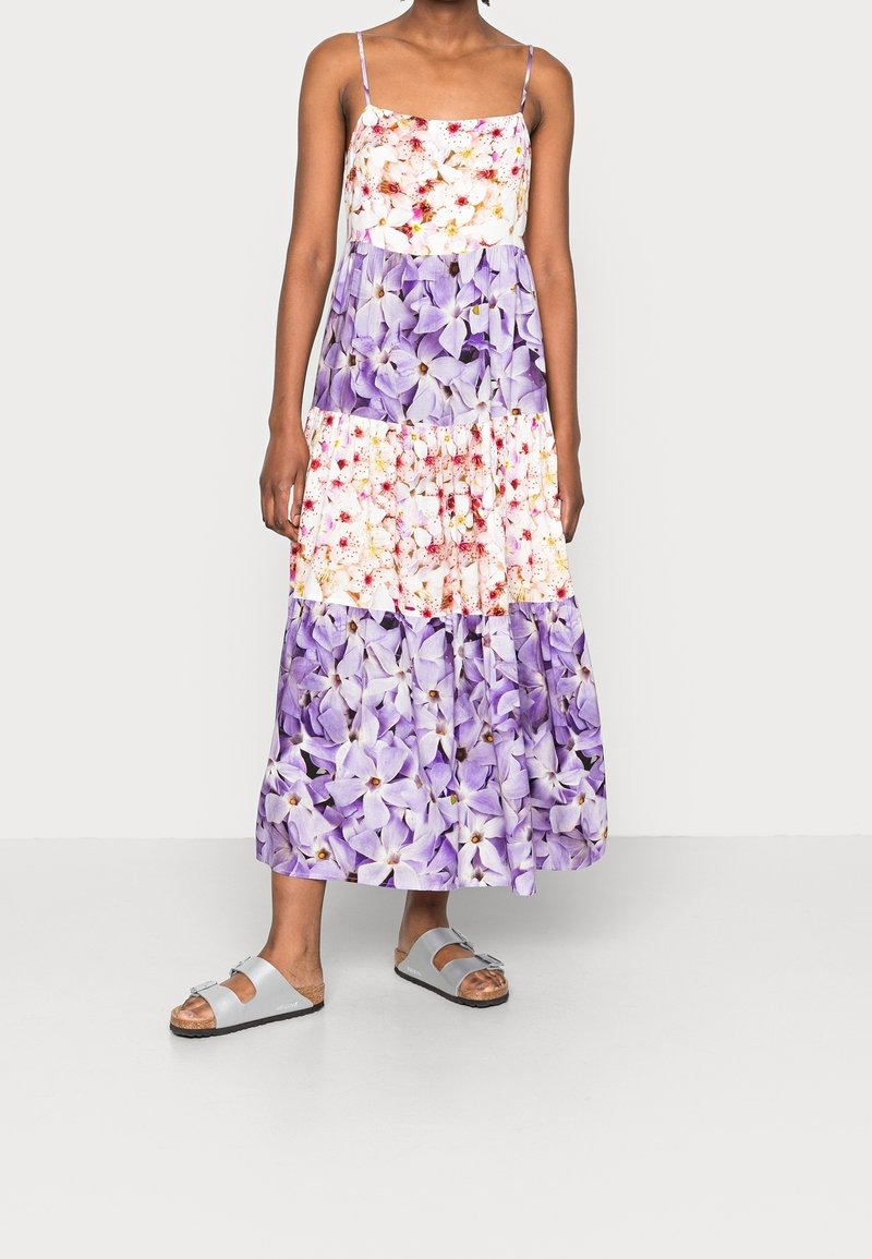 Femme vêtue d'une robe sans manches à volants florale dans des tons de violet et blanc, avec des fines bretelles et des sandales à boucle argentée, se tenant sur un fond uni.