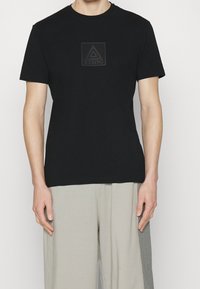 Camiseta de algodón negra con un logotipo rectangular texturizado que presenta un triángulo y el texto "ICEBERG" en el centro, con cuello redondo y mangas cortas.