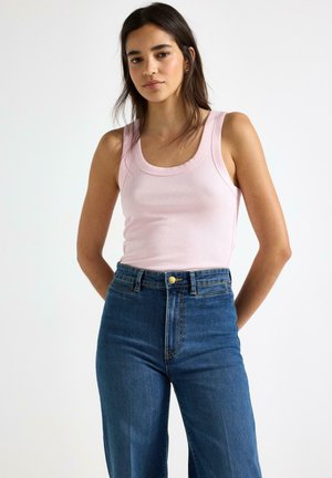 Ung kvinde med langt mørkt hår iført en lyserød ærmeløs top og højttaljede blå jeans, stående foran en ensfarvet hvid baggrund.